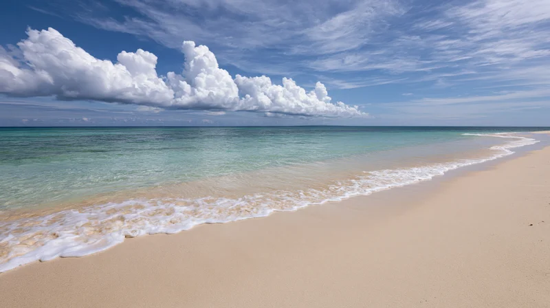 Calm ocean waves rolling onto a flat pale sandy beach under a blue sky with white clouds