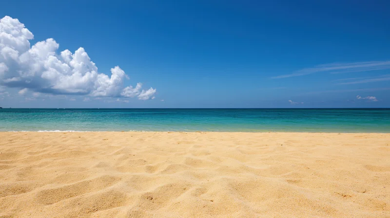 Empty tropical beach with fine golden sand and turquoise ocean under a clear blue sky with scattered clouds