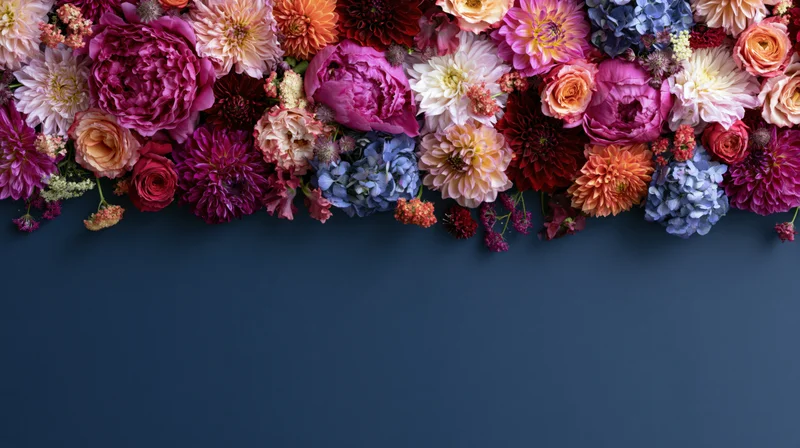 Flat lay of mixed garden flowers including peonies and dahlias arranged along the top of a dark blue surface with copyspace below