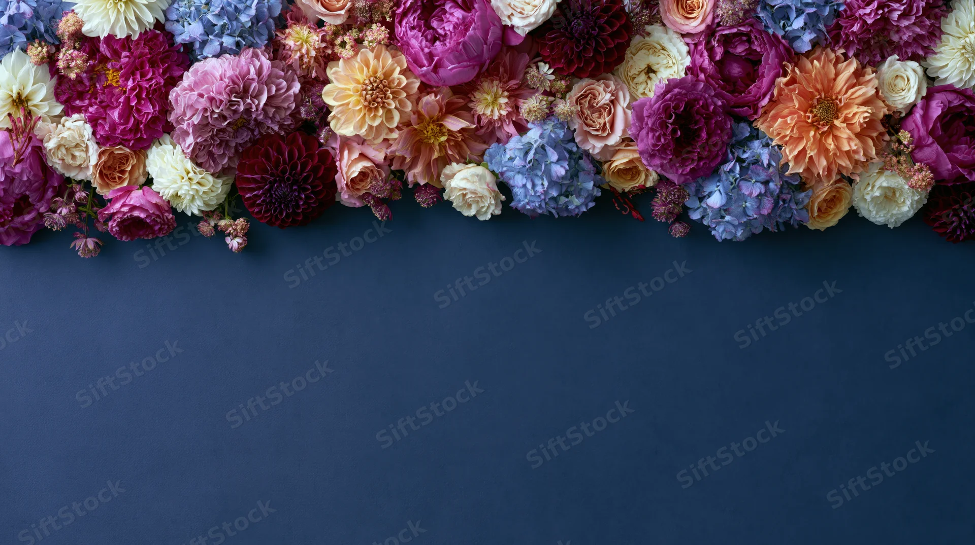 Flat lay of mixed garden flowers including peonies and dahlias arranged along the top of a dark blue surface with copyspace below