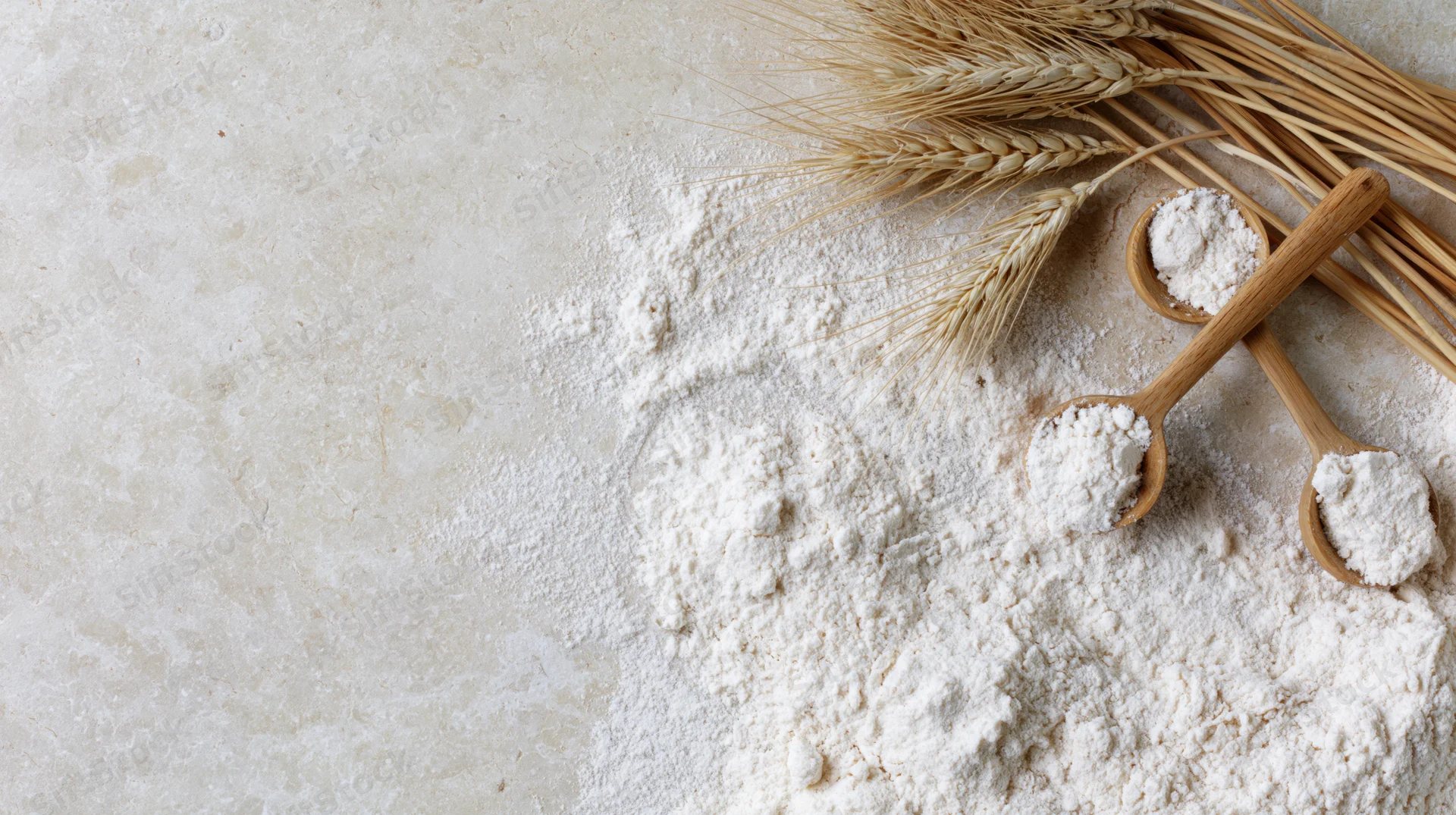 Scattered white flour with wheat stalks and wooden spoons on a cream surface