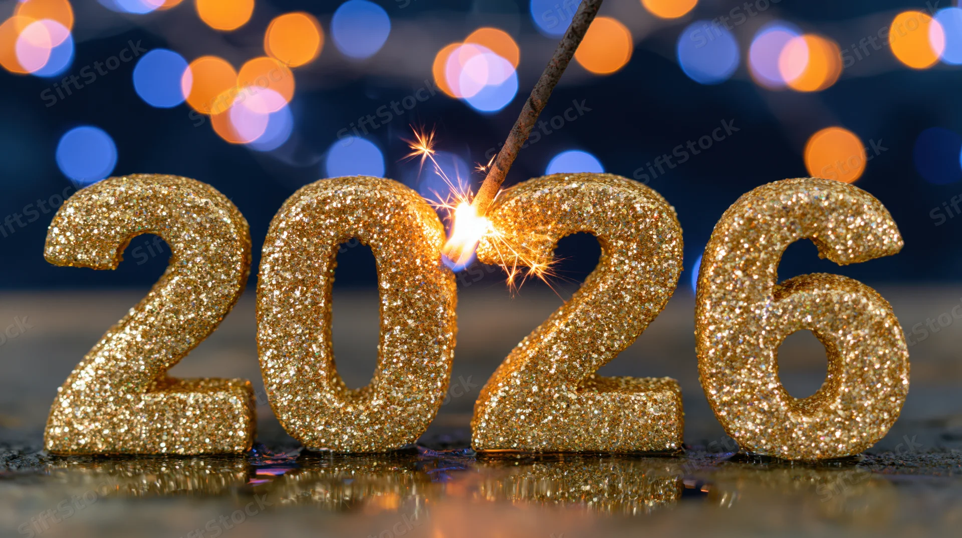 Gold glitter numbers 2026 with sparkler lit on dark reflective surface against bokeh lights