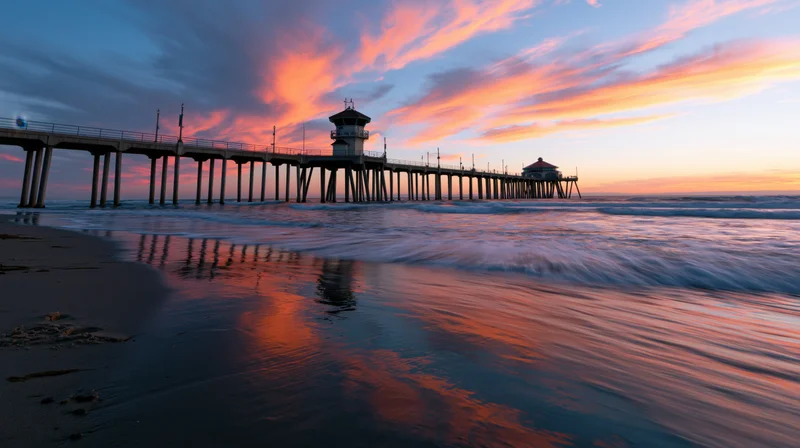 Long pier extending over ocean at sunset with colorful sky reflected in wet sand