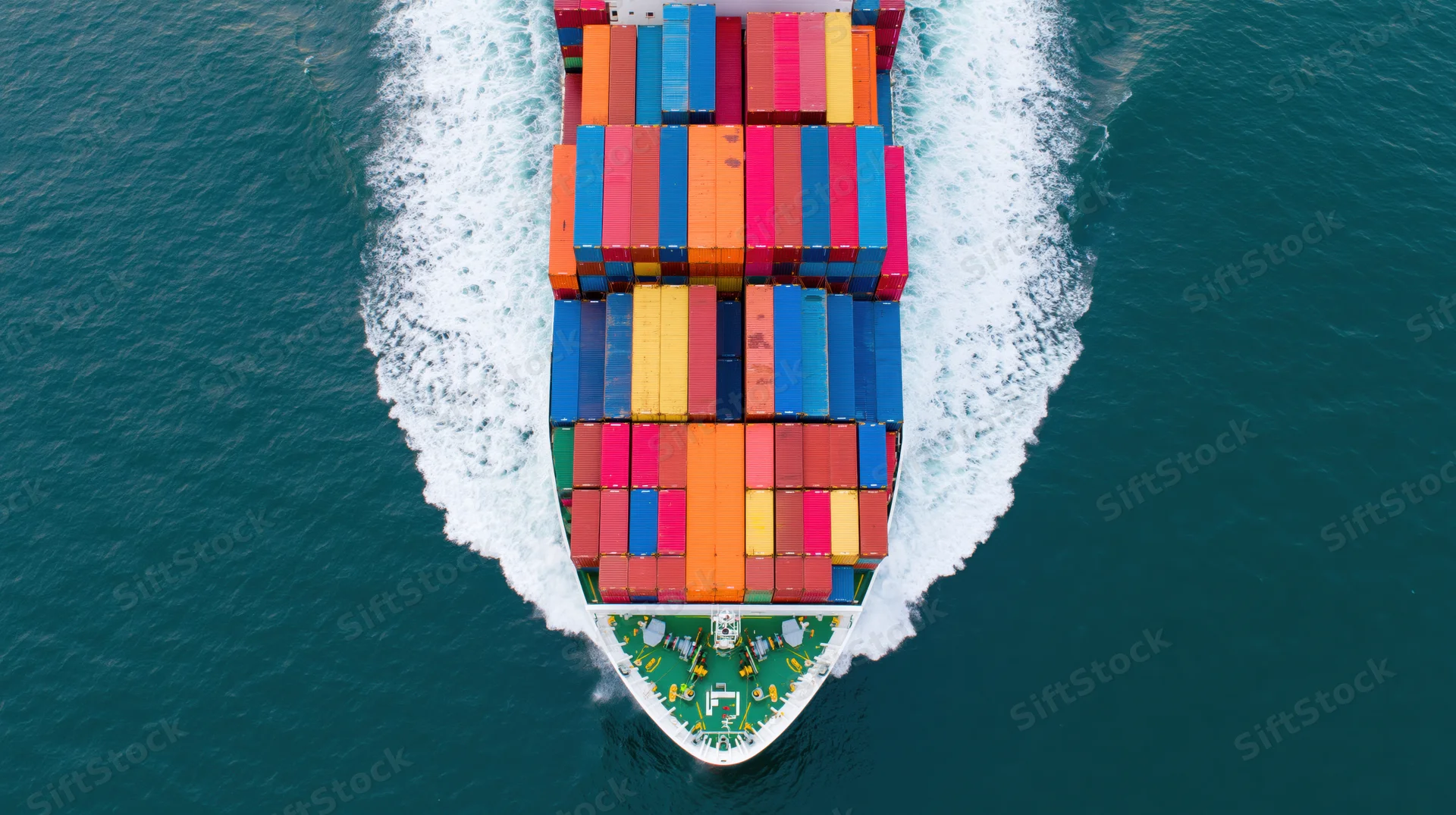 Overhead view of a cargo container ship moving through the ocean with multicolored containers stacked on deck