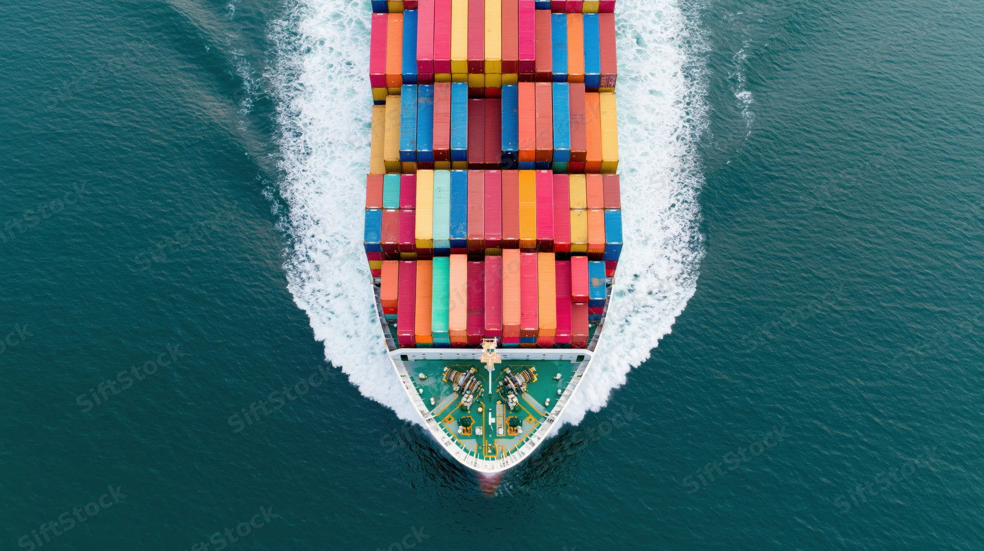Overhead view of a large cargo container ship moving through the ocean with multicolored containers stacked on deck