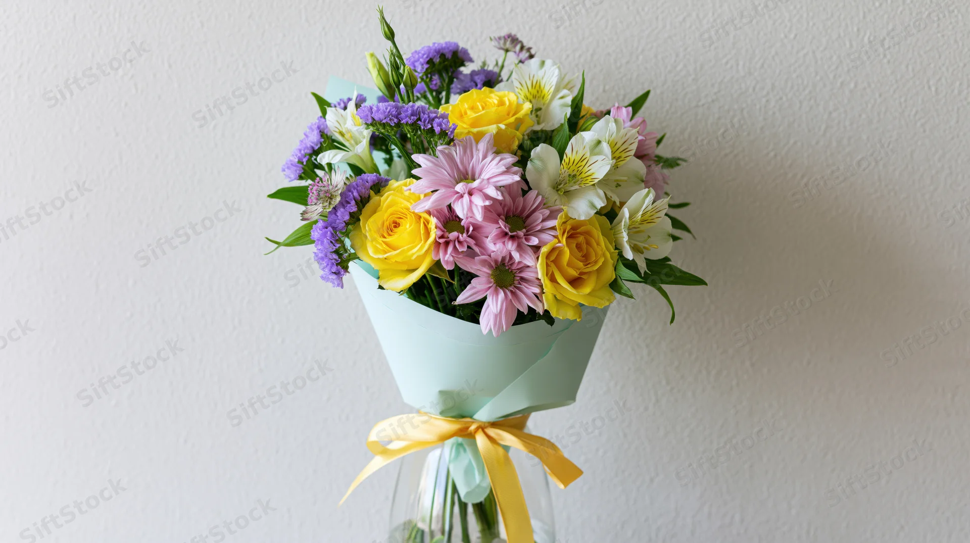 Mixed flower bouquet wrapped in mint paper with yellow ribbon in glass vase against white wall