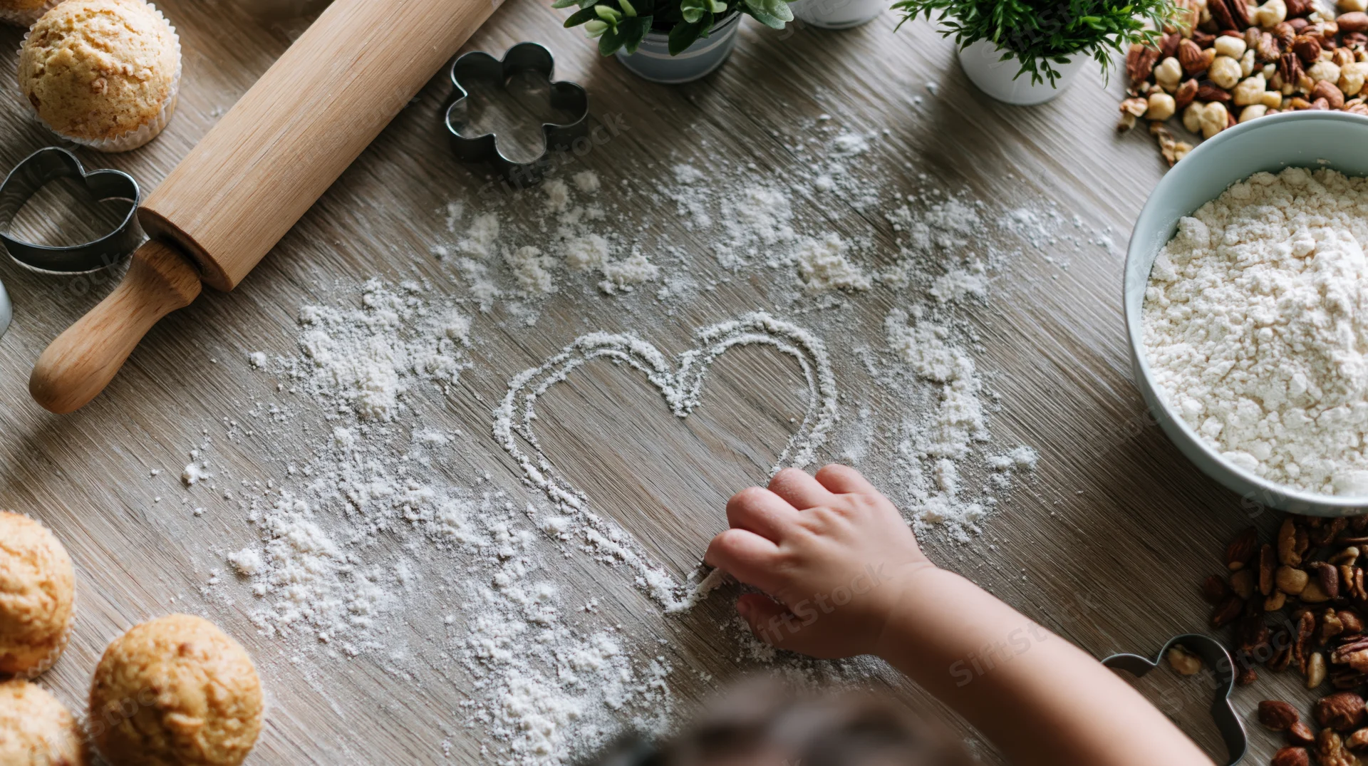 Child’s hand drawing heart in flour on wooden kitchen table surrounded by baking tools and ingredients