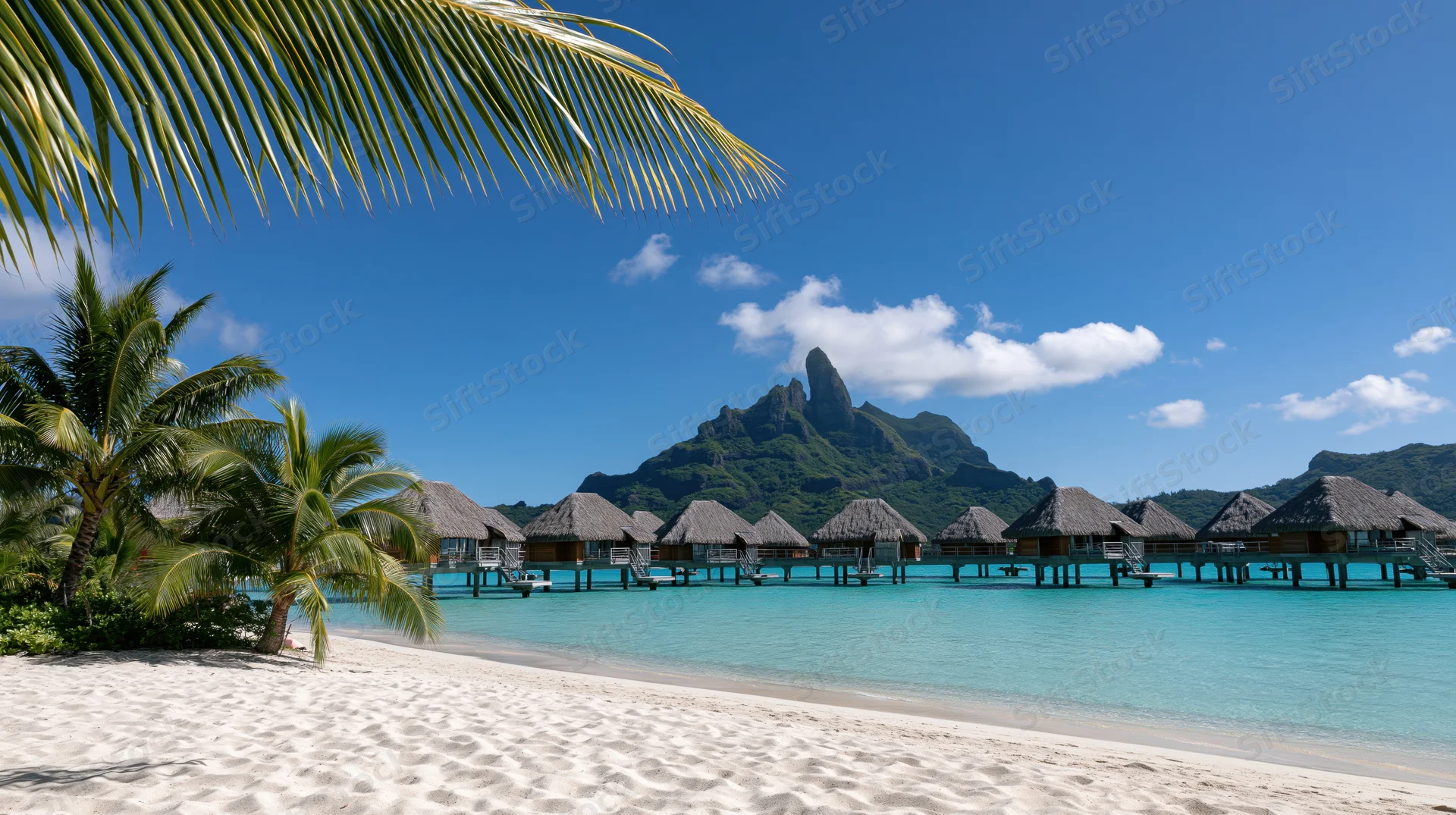 Overwater thatched-roof bungalows on turquoise water beside a sandy beach with palm trees and a mountain in the background