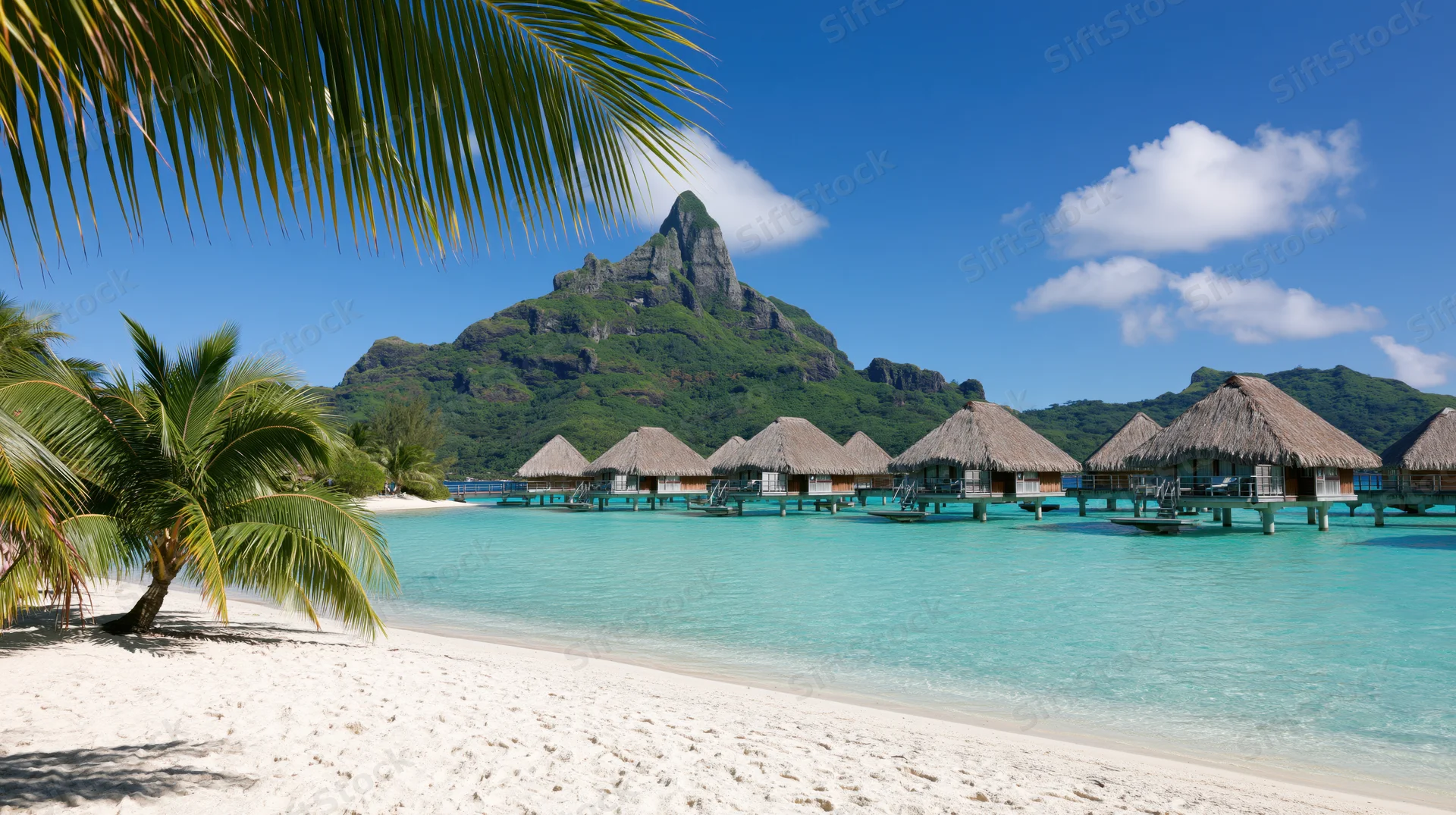 Overwater thatched-roof bungalows on turquoise lagoon beside white sand beach with palm trees and mountain in background