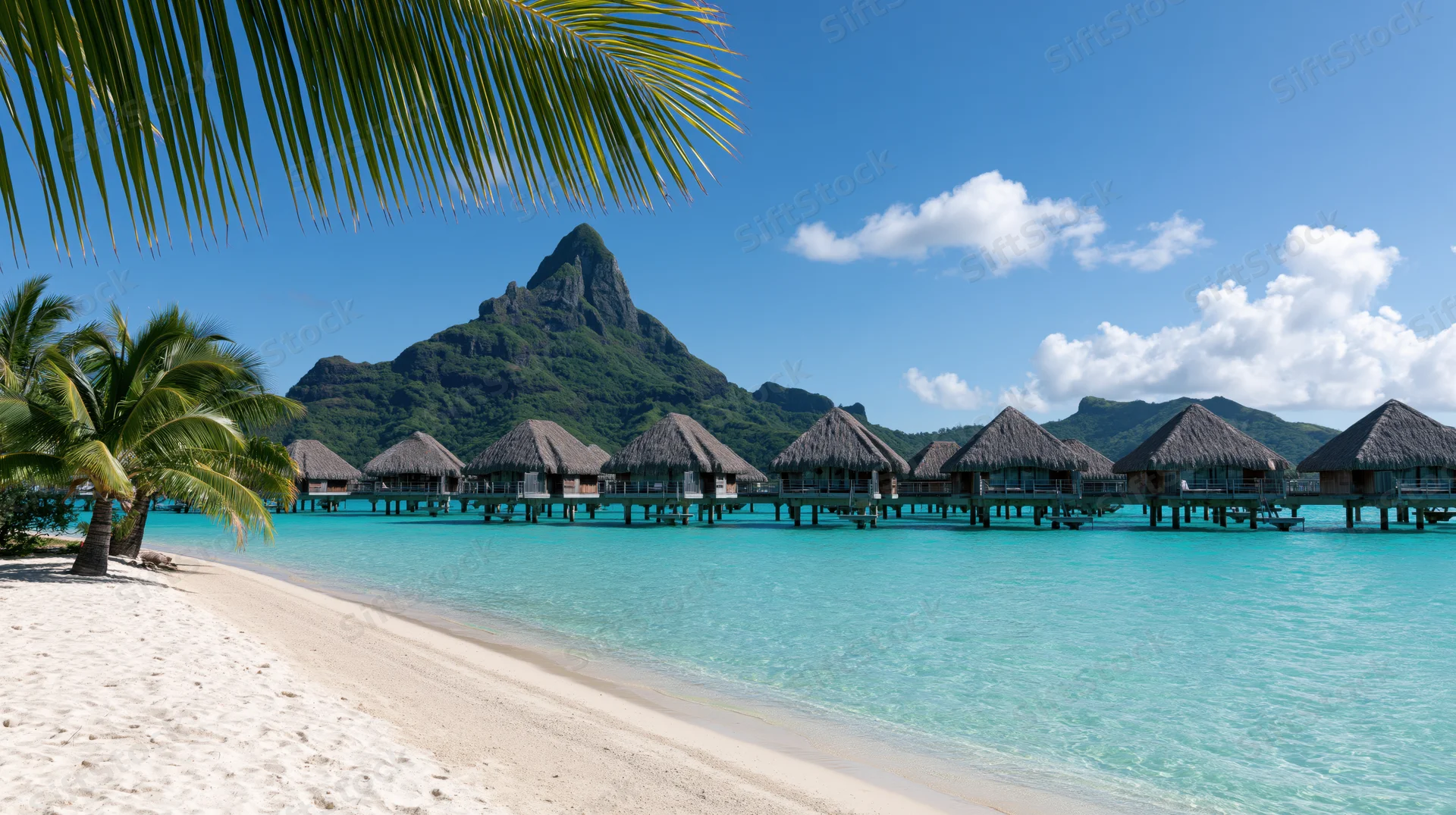 Overwater thatched-roof bungalows on turquoise water beside a white sand beach with palm trees and a green mountain in the background