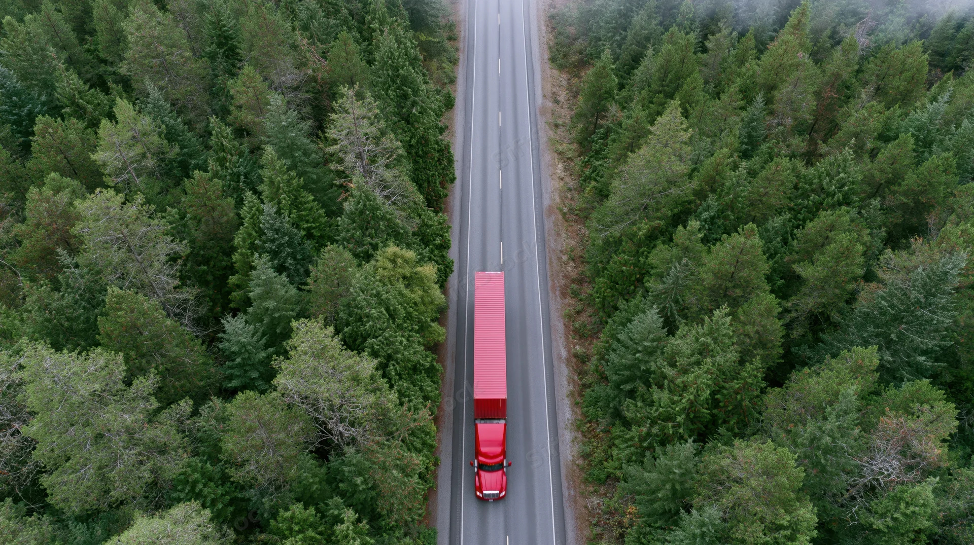 Red semi-truck with full trailer driving along straight highway through dense forest from aerial view