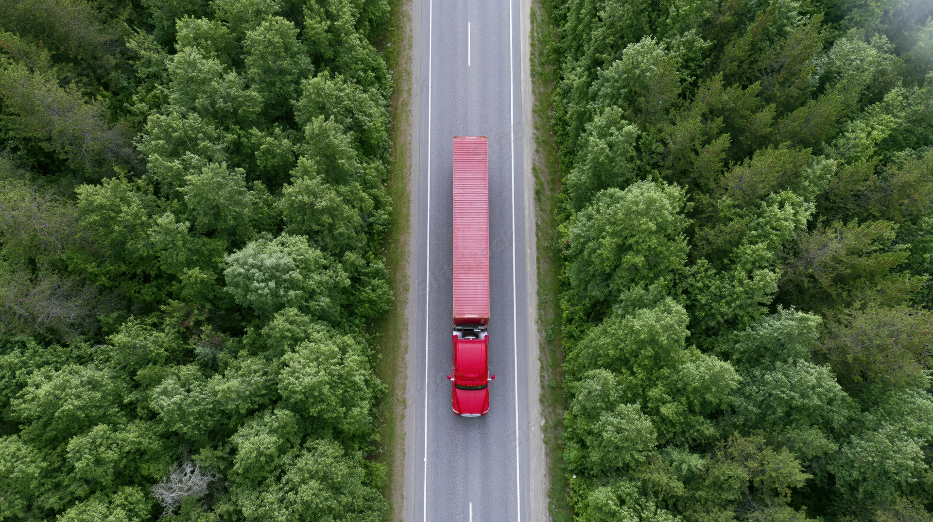 Red semi-truck with full trailer driving straight on a two-lane highway through dense green forest, aerial view