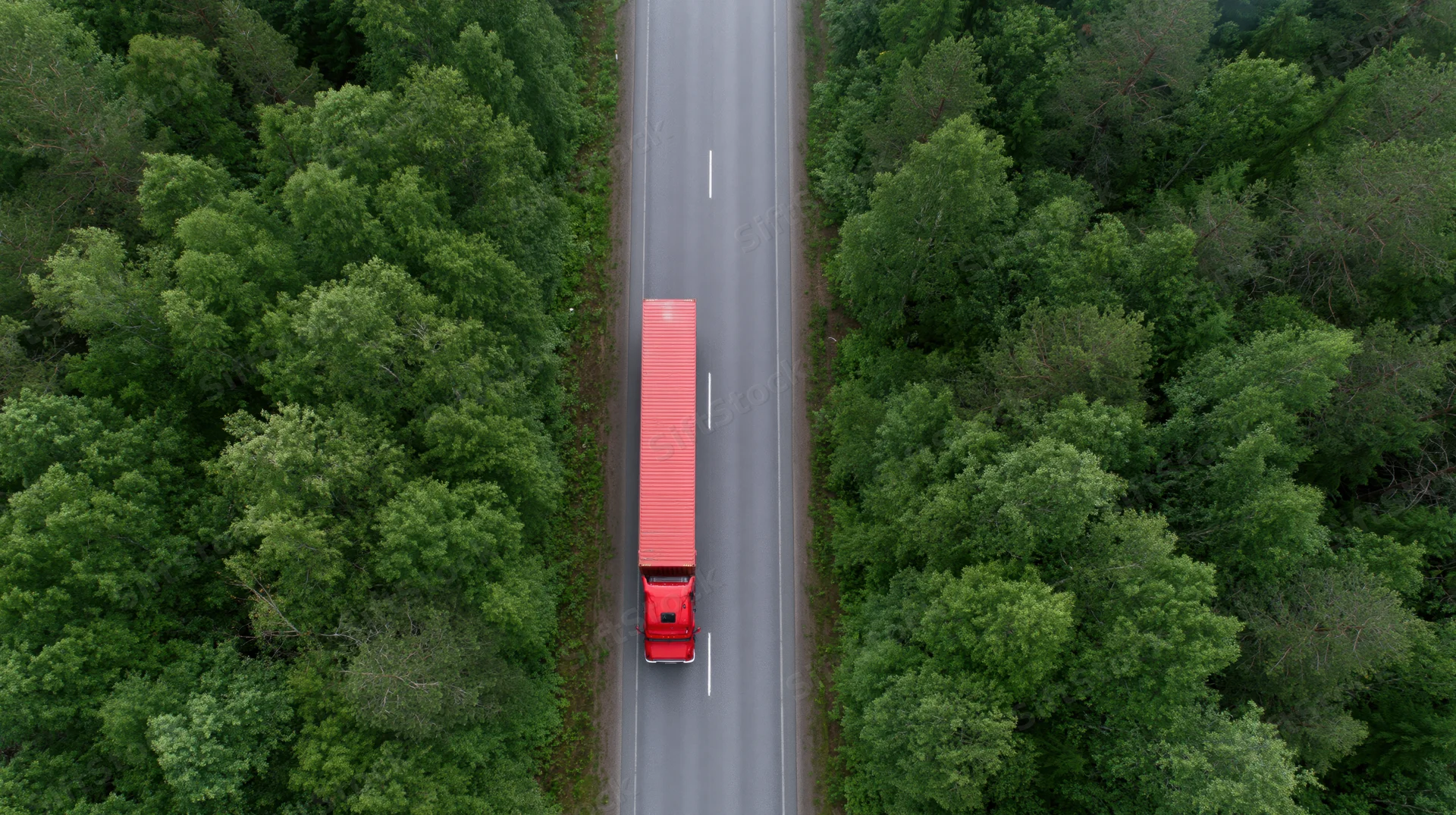 Red semi-truck with full trailer driving straight on a two-lane highway through dense green forest, aerial top-down view