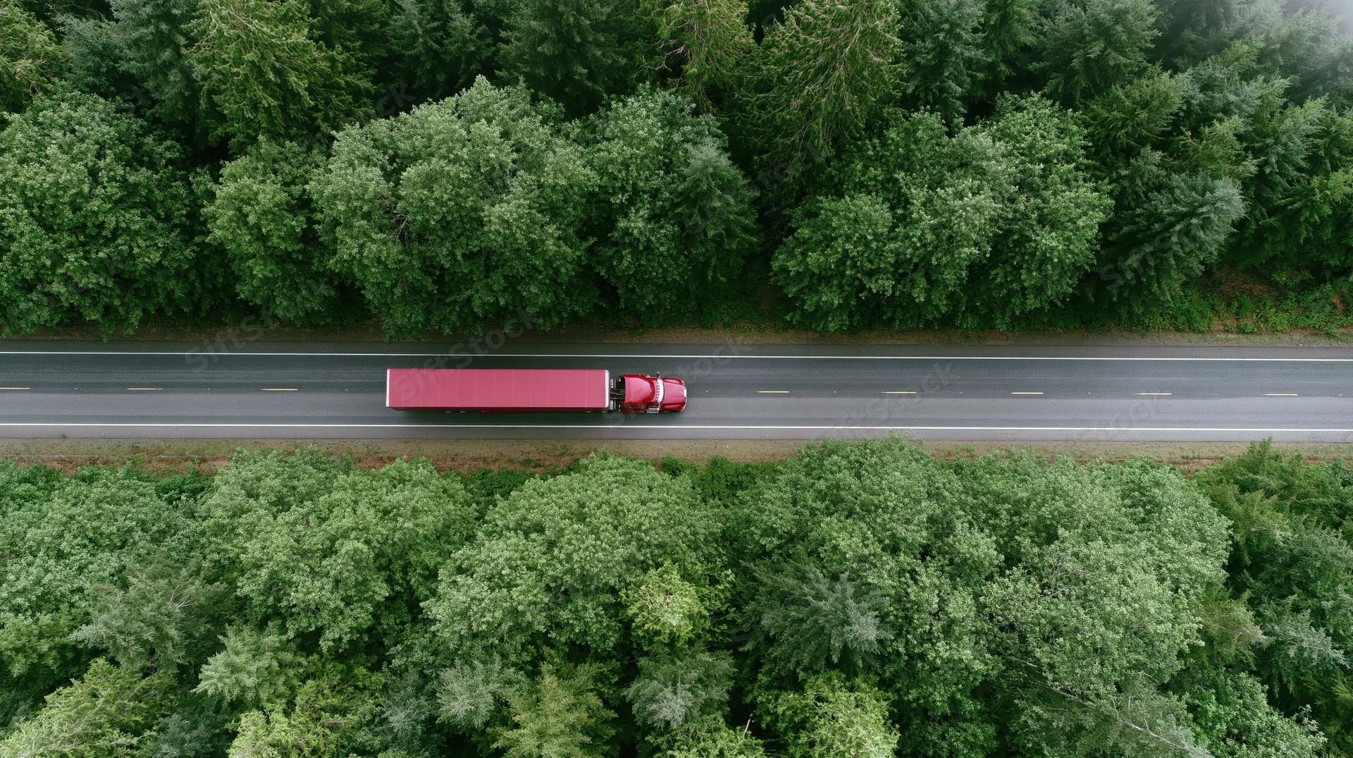 Red semi-truck with full trailer driving along straight highway surrounded by dense green forest, aerial view