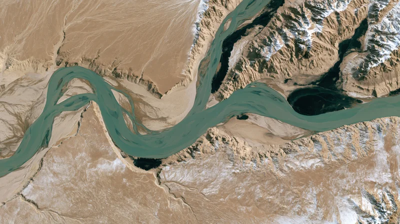 Satellite view of a river delta winding through arid terrain with snow-capped mountains and canyons