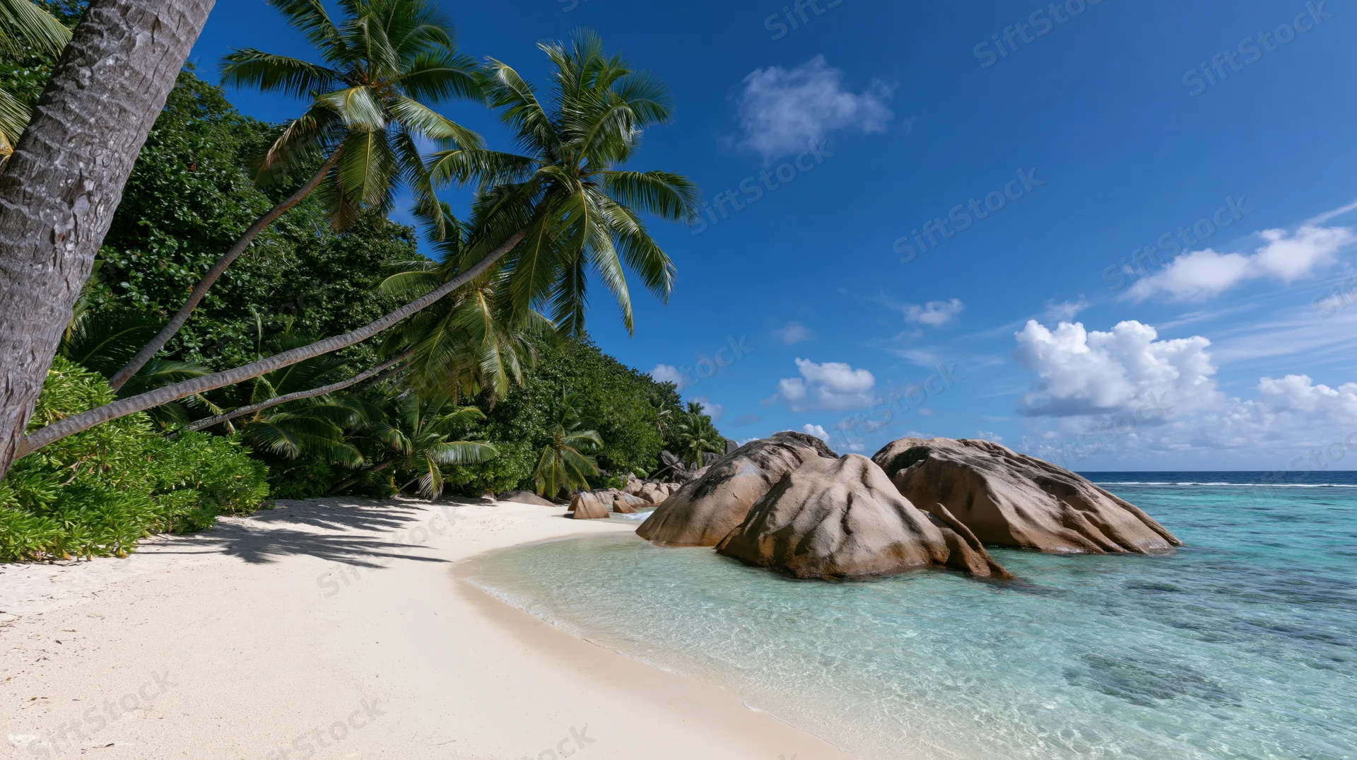 Secluded tropical beach with white sand curving toward large rocks and palm trees under blue sky