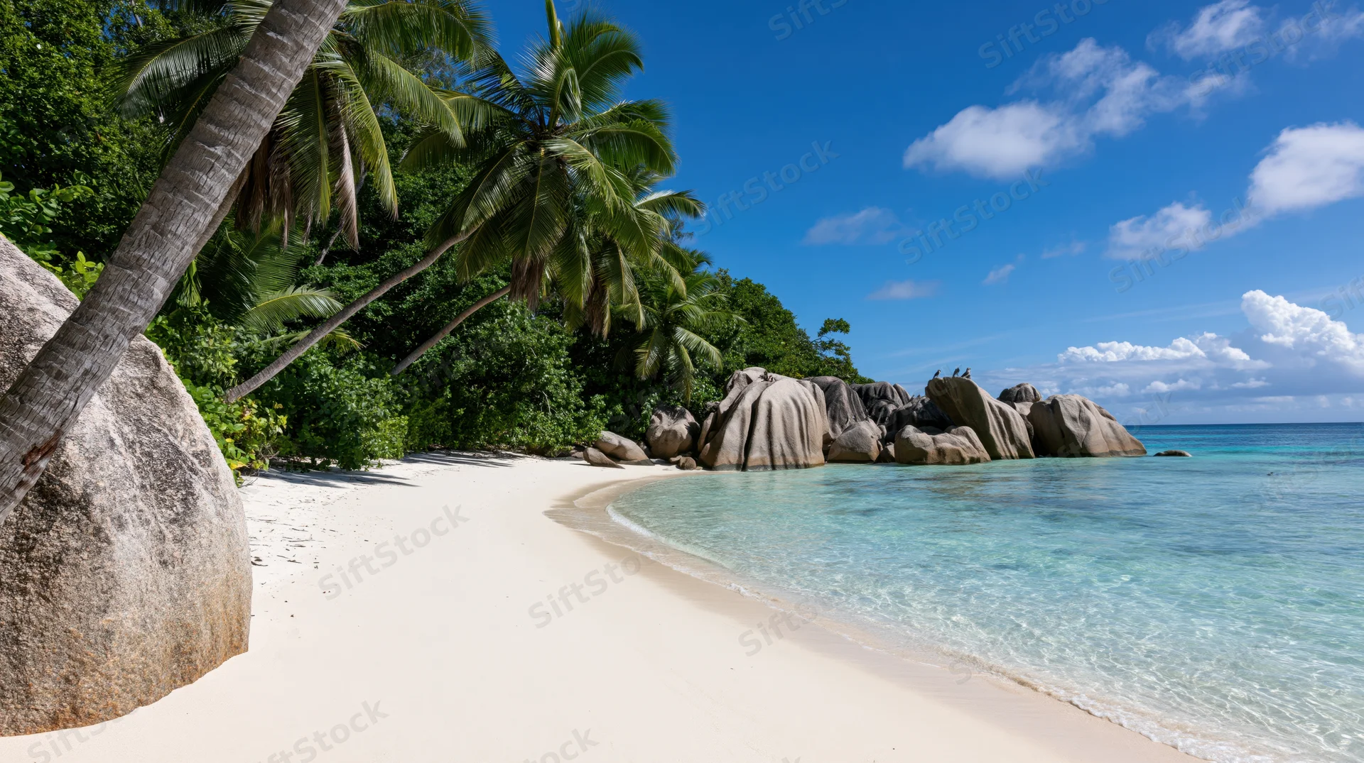 Secluded tropical beach with white sand curving toward large rocks and palm trees under blue sky