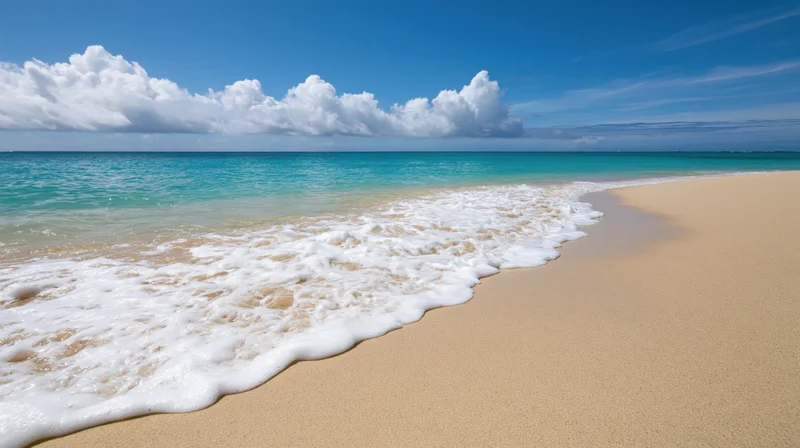 Shallow ocean wave washing gently onto a flat sandy beach under a blue sky with white clouds