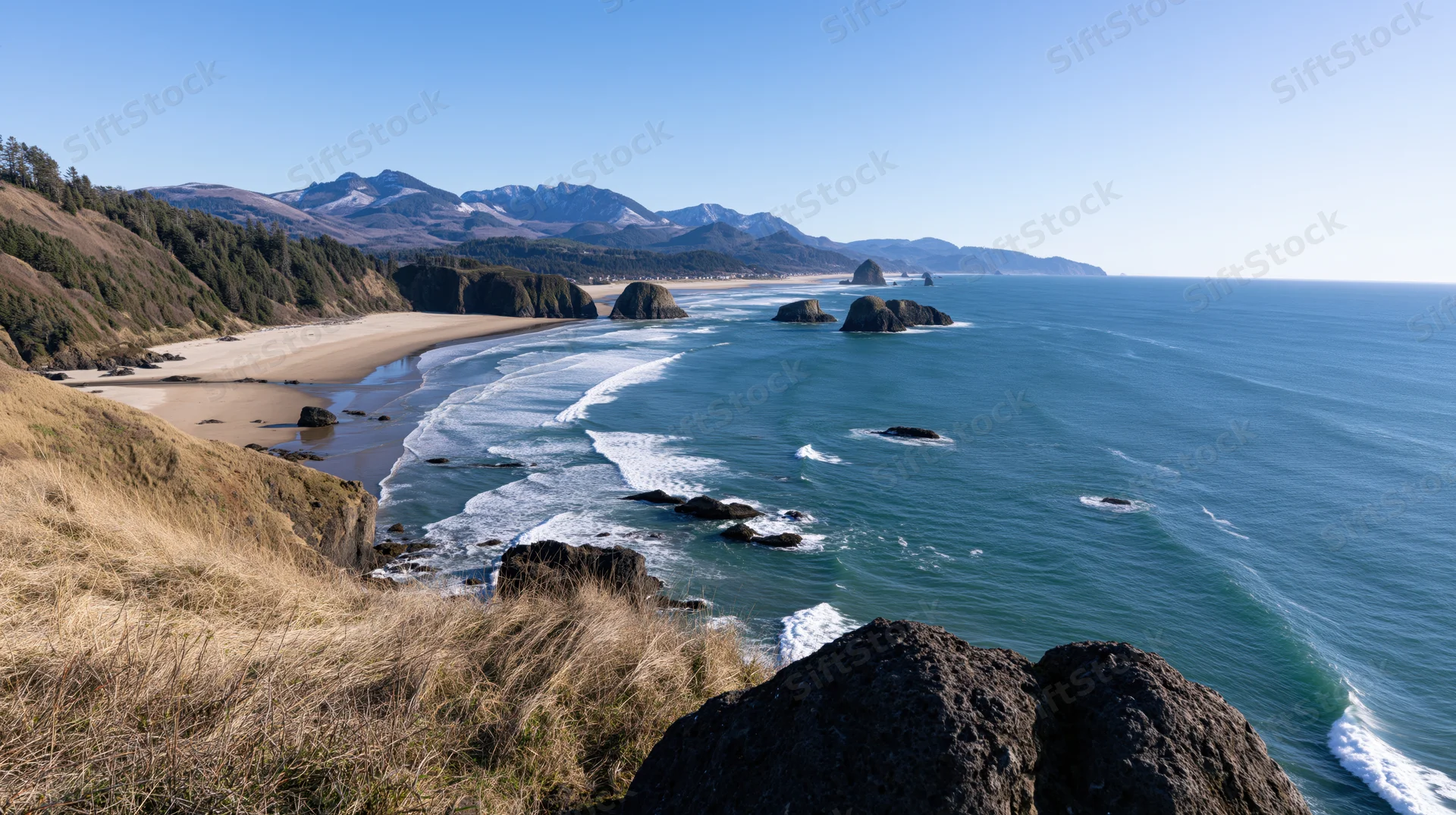 Sweeping view of a curved pacific northwest coastline with waves breaking on sandy beach and rocky cliffs under clear blue sky