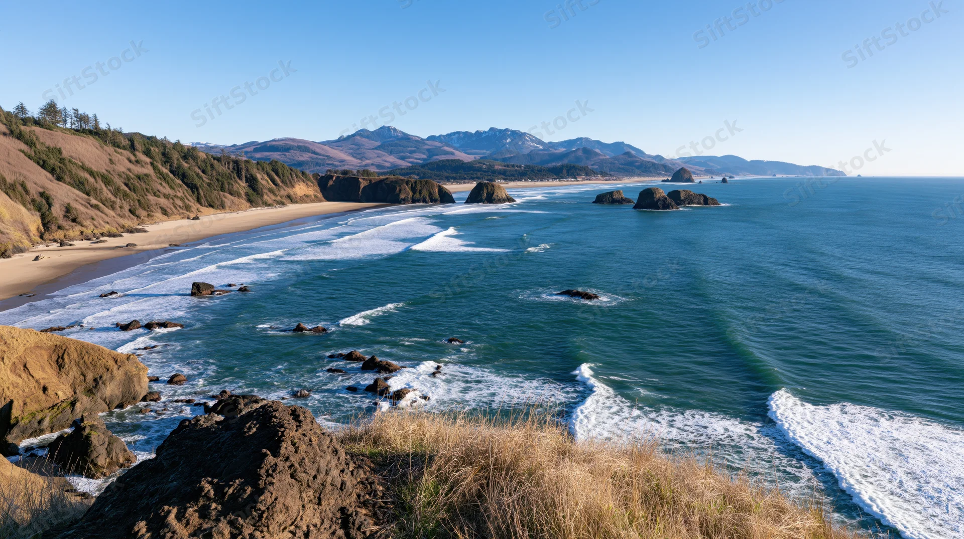 Sweeping view of a curved pacific northwest coastline with waves breaking on sandy beach and rocky cliffs under clear blue sky