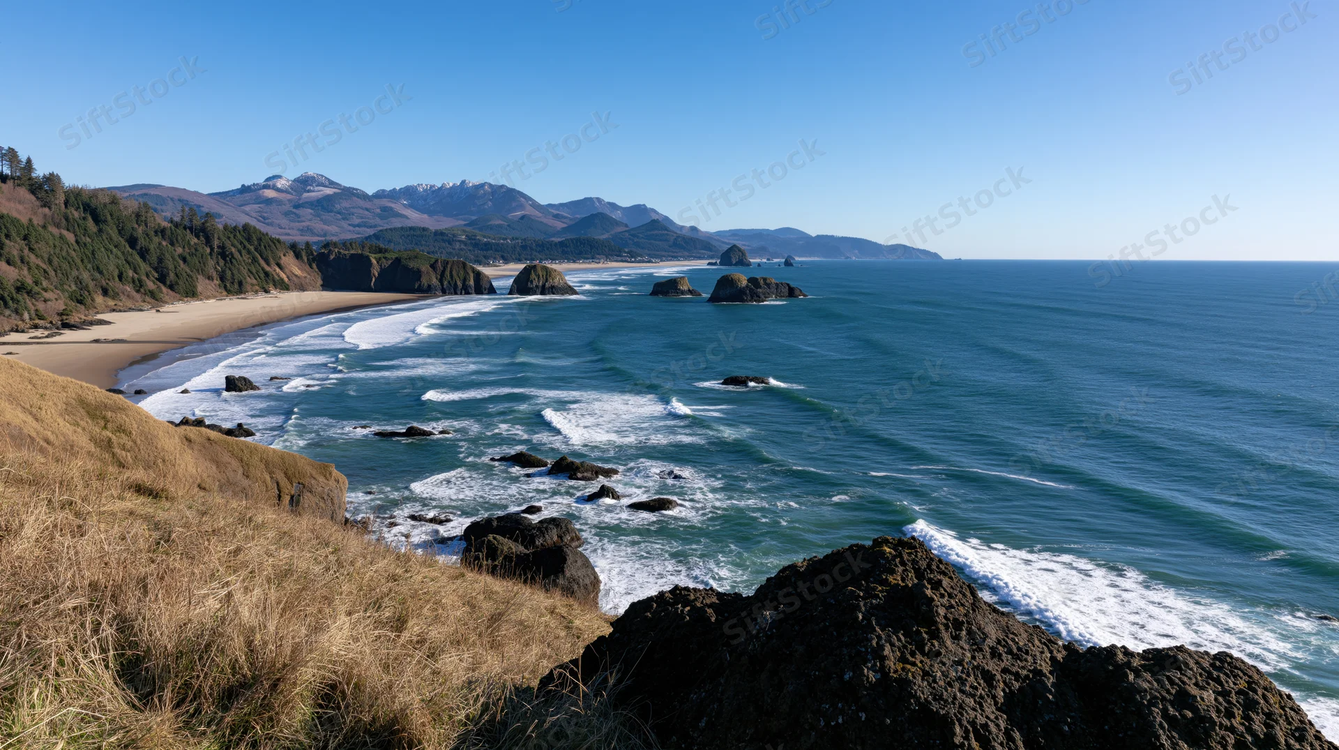 Sweeping aerial view of a curved pacific northwest coastline with waves breaking on rocky shore and distant mountains under clear blue sky