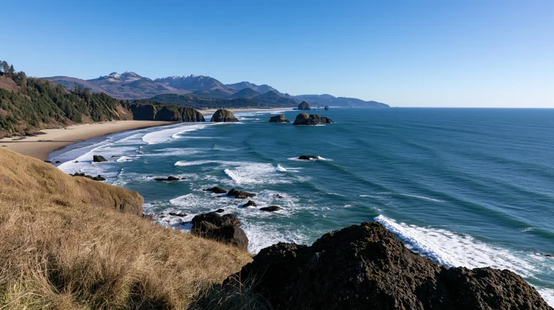 Sweeping aerial view of a curved pacific northwest coastline with waves breaking on rocky shore and distant mountains under clear blue sky