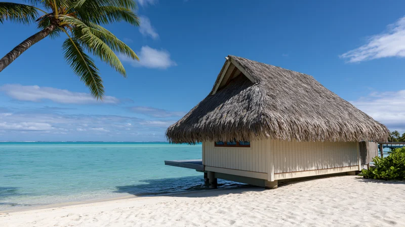 Thatched-roof overwater bungalow on sandy beach beside turquoise ocean under blue sky