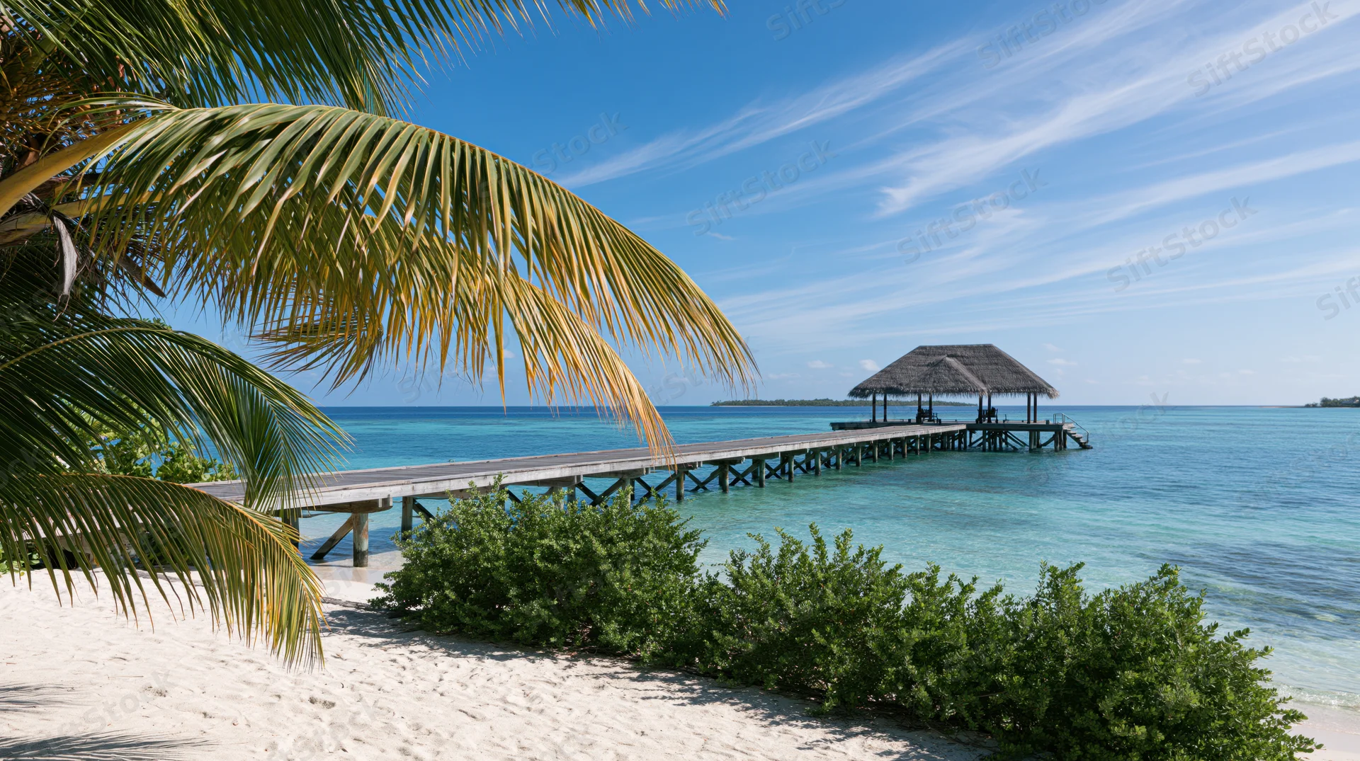 Wooden pier extending over turquoise ocean from sandy beach with palm trees and thatched hut at end