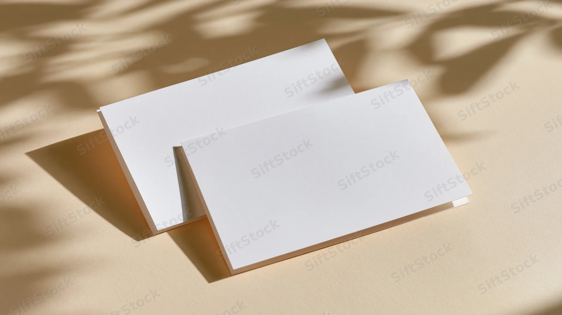 Two blank white cards overlapping on a beige surface with leaf shadows from sunlight