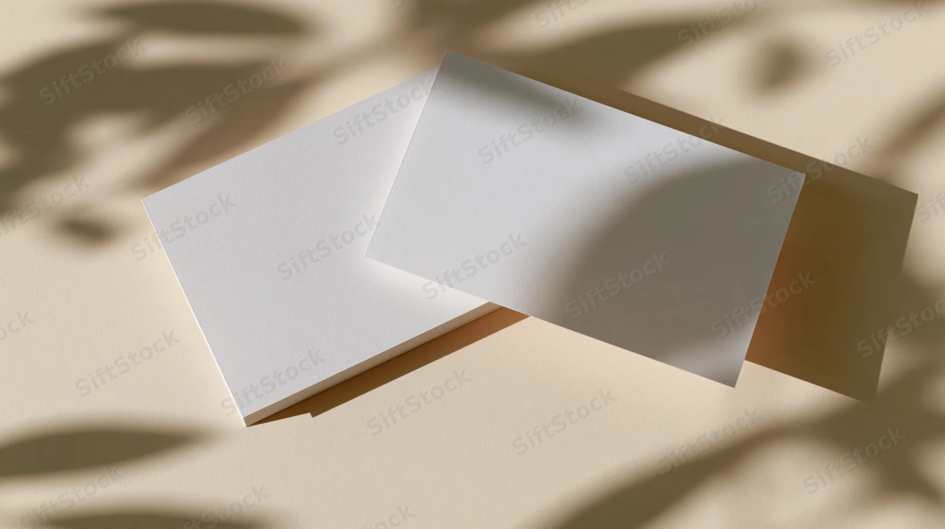 Two blank white rectangular cards overlapping slightly on a beige surface with leaf shadows from sunlight