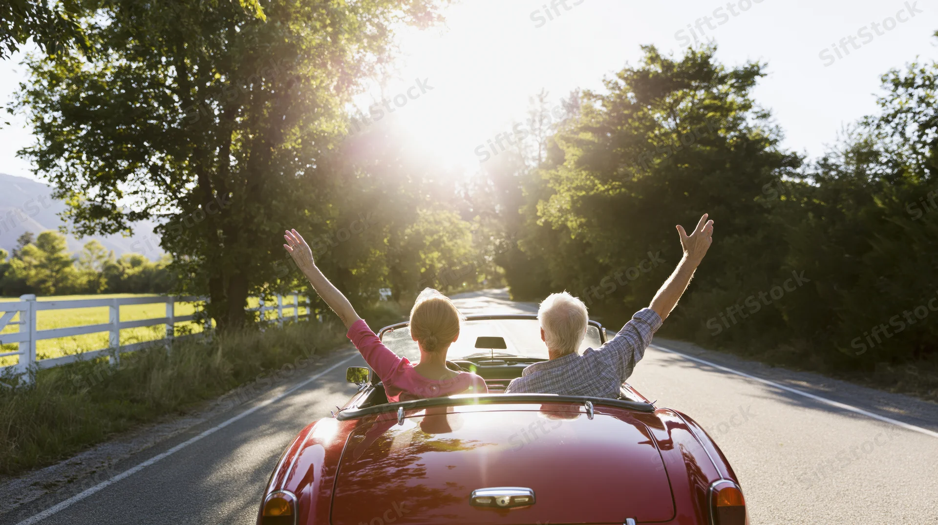 Senior couple driving red classic convertible on country road with arms raised toward sunset