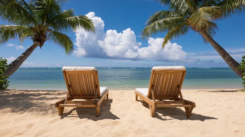 Two wooden beach loungers on pale golden sand framed by palm trees overlooking ocean under blue sky with white clouds
