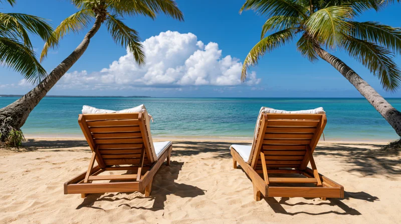 Two empty wooden beach loungers on pale golden sand framed by palm trees overlooking turquoise ocean under blue sky with white clouds