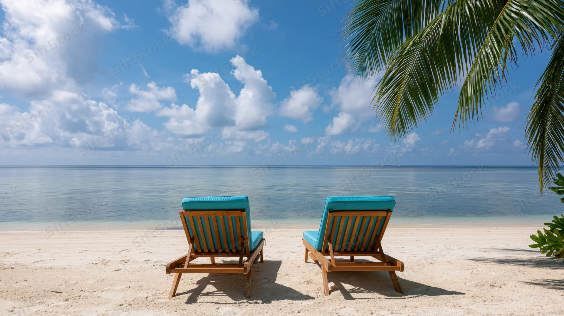 Two empty wooden beach loungers with turquoise cushions facing the ocean on a sandy shore under a blue sky with clouds