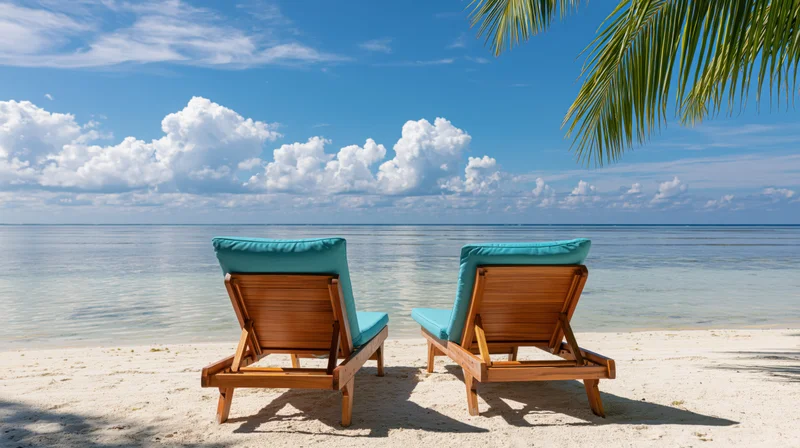 Two wooden beach loungers with turquoise cushions facing the ocean on a sandy shore under a palm tree