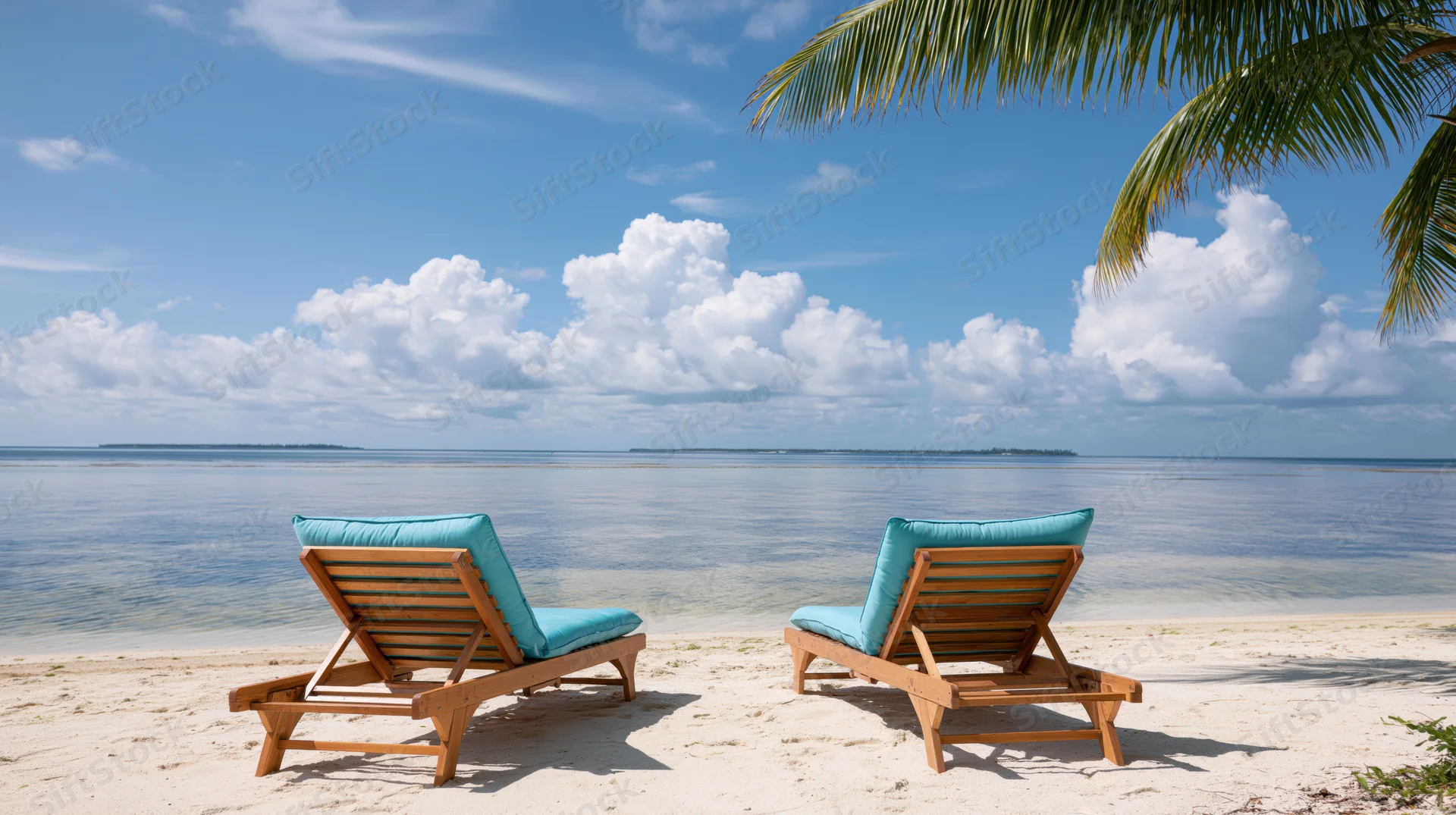 Two empty wooden beach loungers with turquoise cushions on a sandy shore facing the ocean under a blue sky with clouds and palm fronds overhead