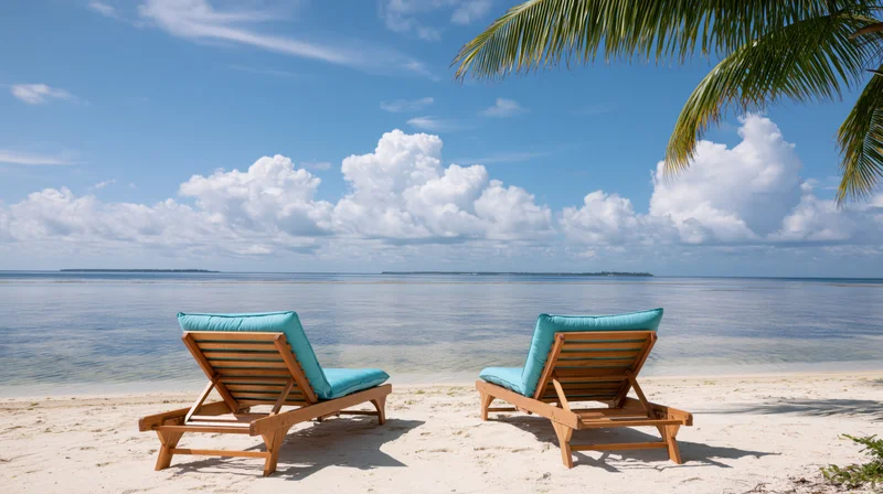 Two empty wooden beach loungers with turquoise cushions on a sandy shore facing the ocean under a blue sky with clouds and palm fronds overhead