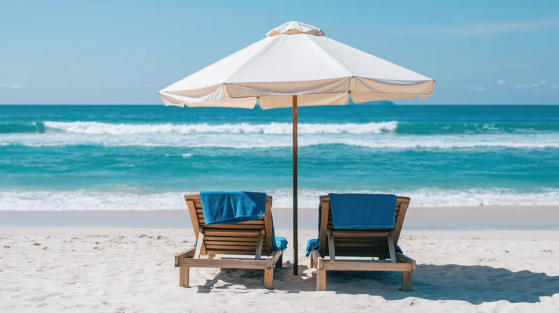 Two wooden beach loungers with blue towels under a white umbrella on sandy shore facing ocean waves
