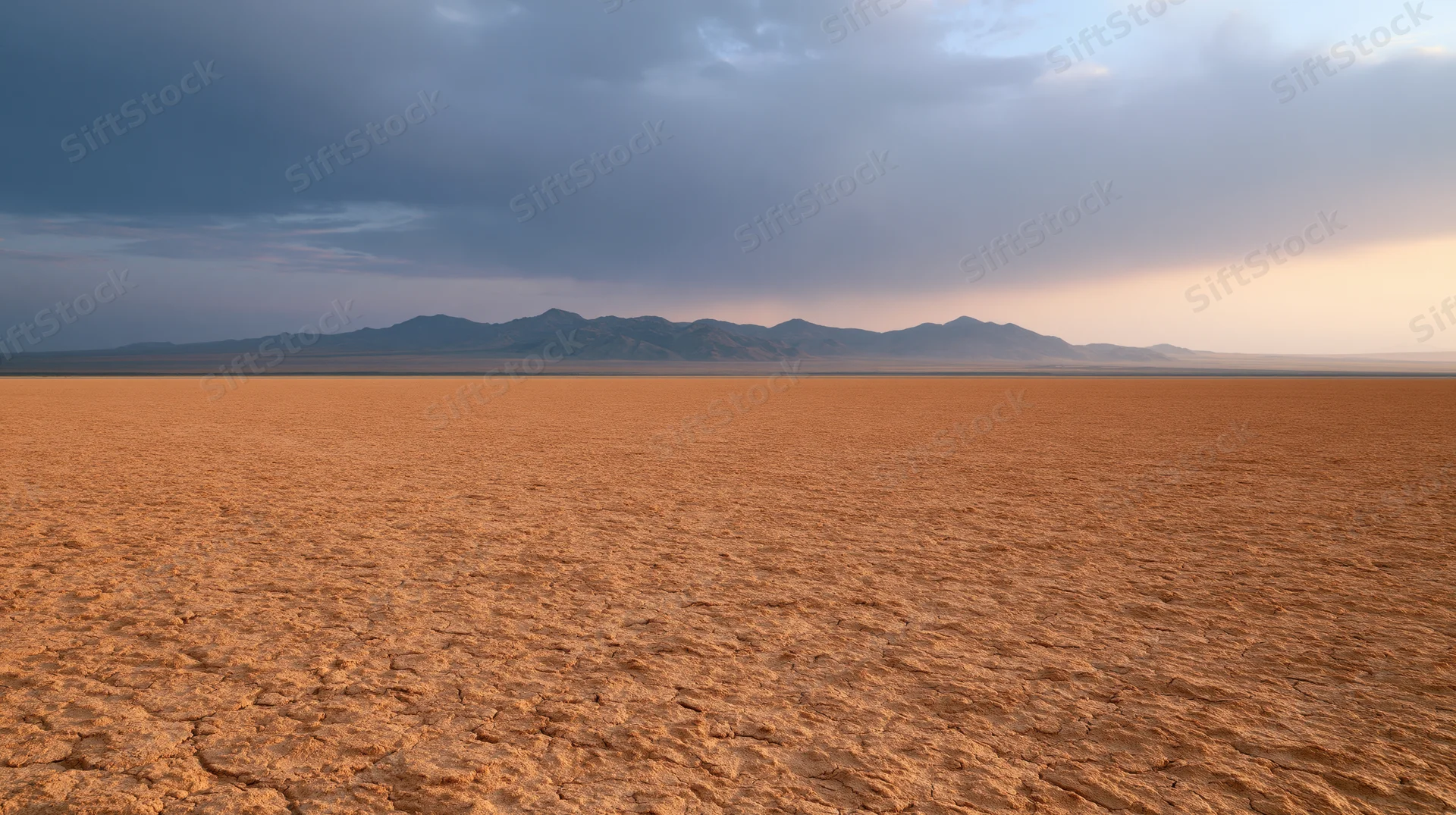 Vast cracked desert plain stretching toward distant mountains under overcast sky at dusk