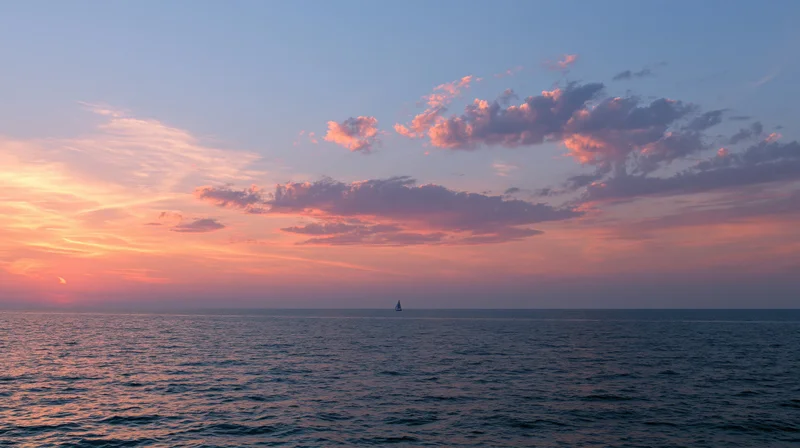 Sailboat silhouetted on calm ocean at sunset under pink and orange sky