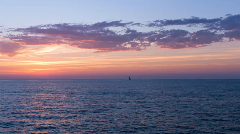 Distant sailboat silhouetted on calm ocean at sunset under colorful sky