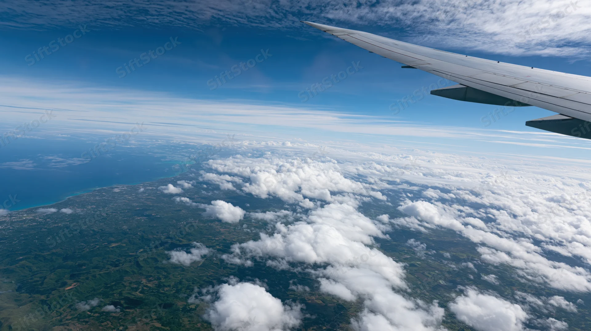 Airplane wing extending over clouds and green landscape below from a window view during daytime flight
