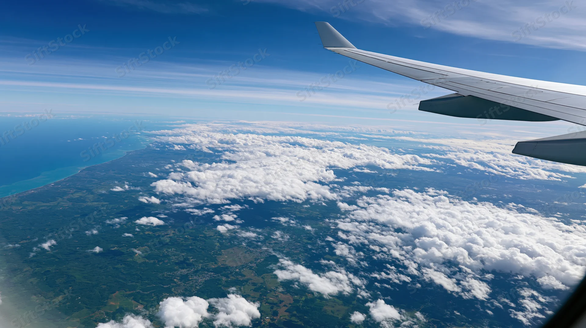 Airplane wing visible through window overlooking clouds and coastline below during daytime flight