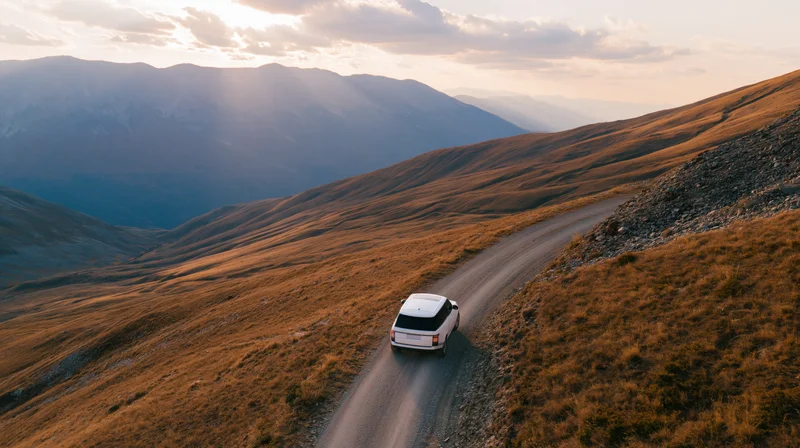 White suv driving away on winding gravel road through mountain valley at sunset