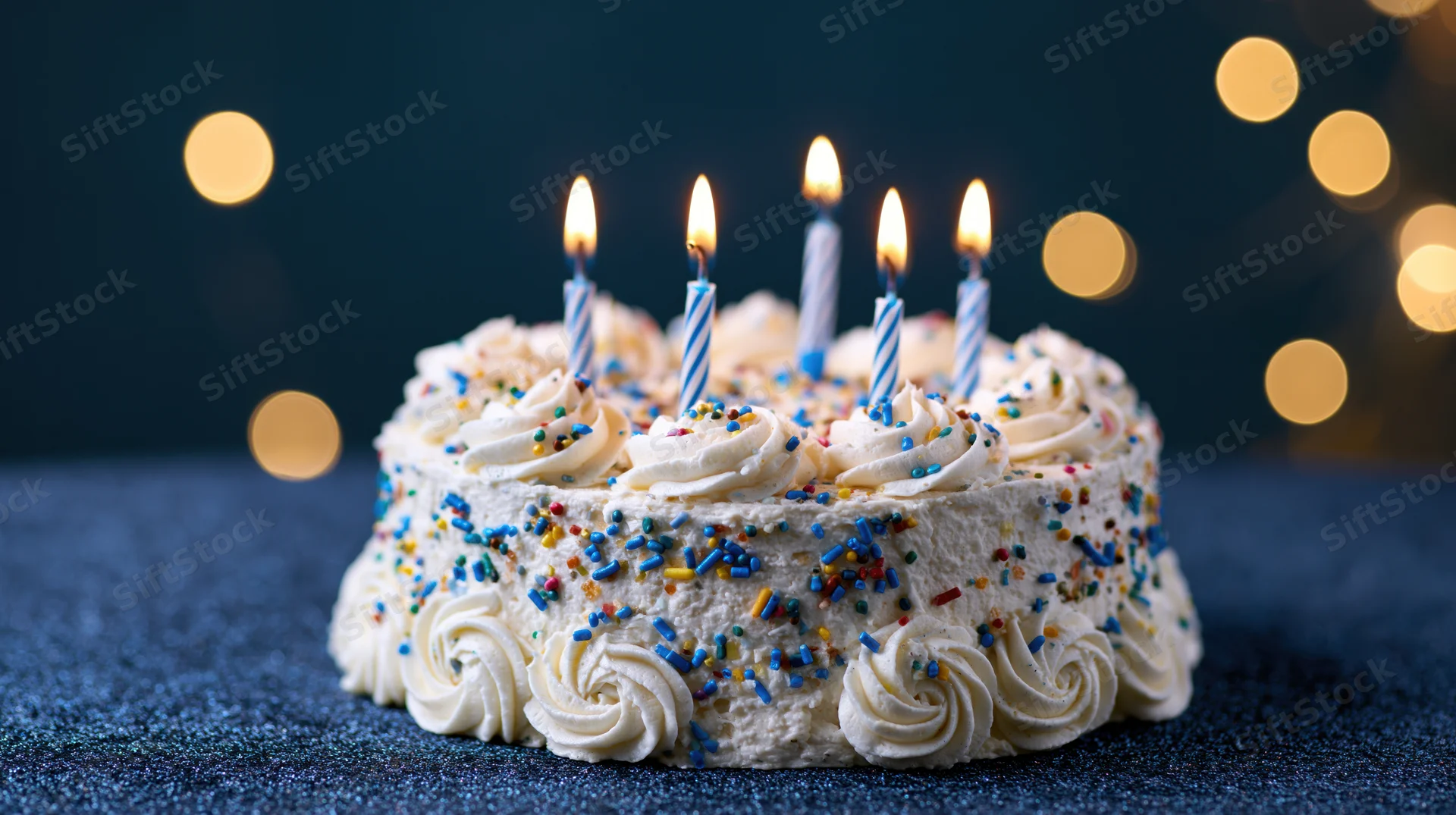 White frosted birthday cake with lit candles and colorful sprinkles on dark surface