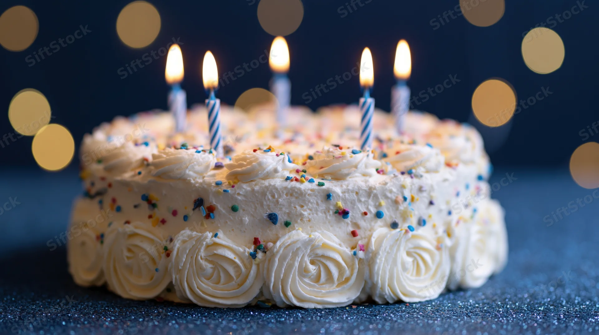 White frosted birthday cake with lit candles and colorful sprinkles on a dark glittery surface with bokeh lights in the background