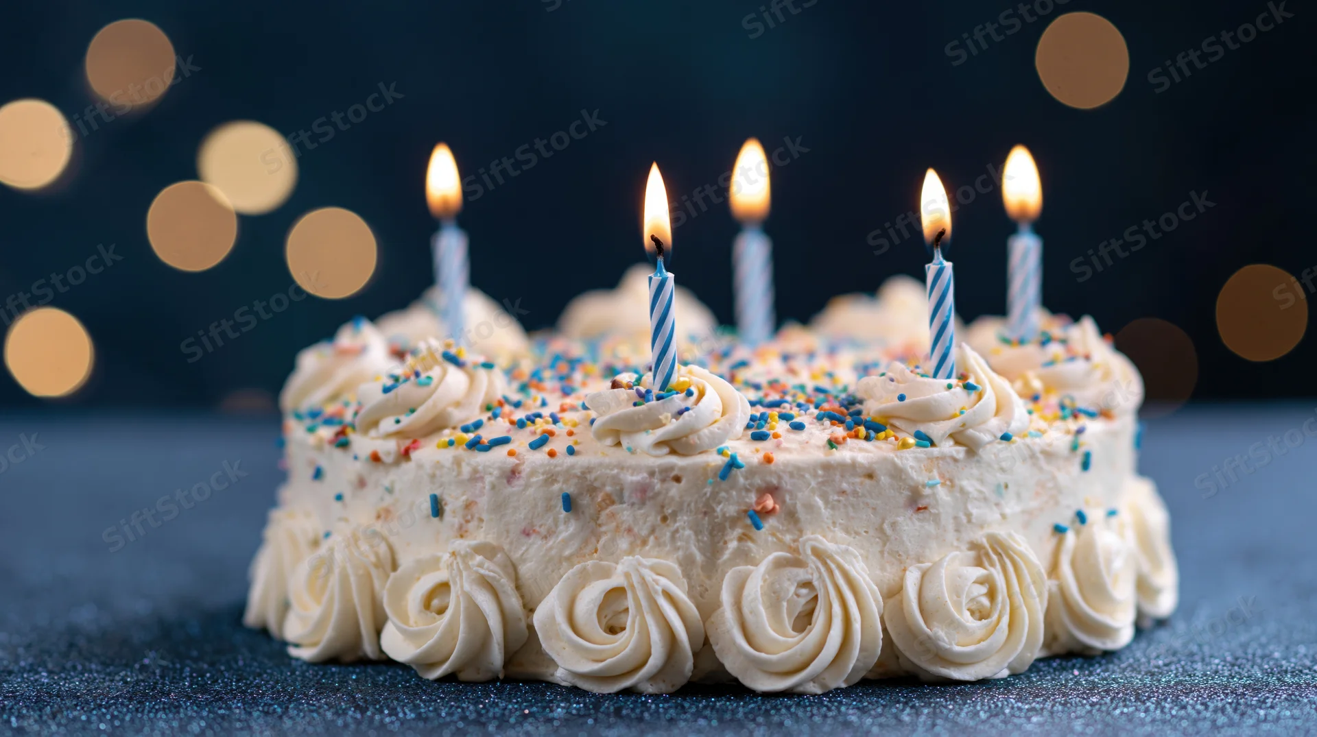 White frosted birthday cake with lit candles and colorful sprinkles on a dark surface
