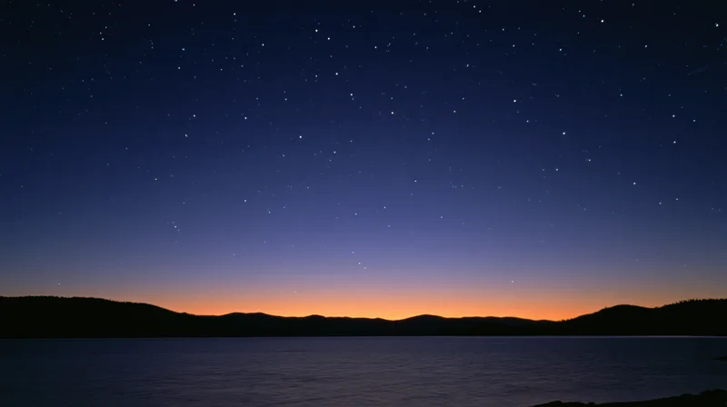 Starry night sky over calm lake with distant mountains at twilight