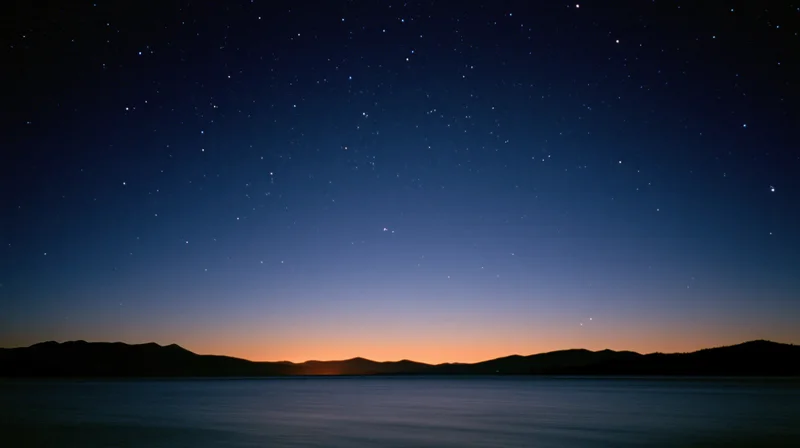 Starry night sky over calm lake with silhouetted mountains at twilight