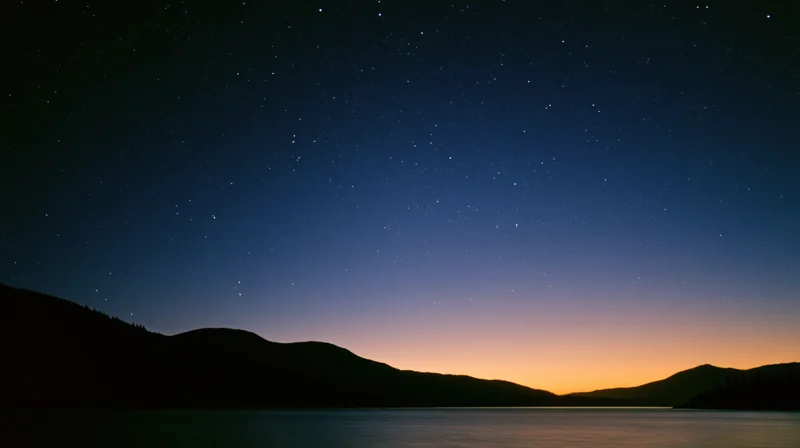 Starry night sky over calm lake with mountain silhouette at twilight